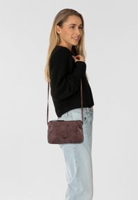 Borsa a tracolla compatta in materiale mauve texturizzato, con chiusura a zip e tracolla regolabile, abbinata a jeans azzurri chiari e a un maglione nero.