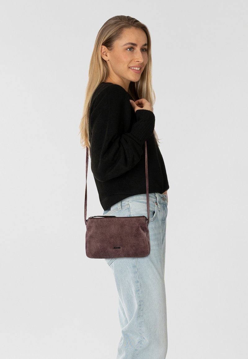 Borsa a tracolla compatta in materiale mauve texturizzato, con chiusura a zip e tracolla regolabile, abbinata a jeans azzurri chiari e a un maglione nero.