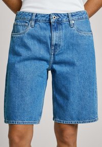 Persona que lleva pantalones cortos de mezclilla azules de media longitud con botón frontal y bolsillos, combinados con una blusa sin mangas blanca.