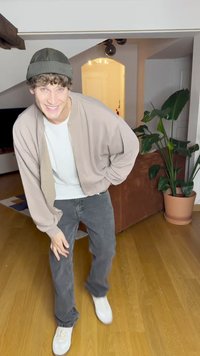 Μπεζ ζακέτα bomber με φερμουάρ πάνω από ένα λευκό T-shirt, συνδυασμένη με σκούρα γκρι τζιν και λευκά αθλητικά παπούτσια. Ένα πράσινο πλεκτό σκουφάκι ολοκληρώνει την εμφάνιση.