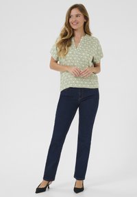 Blusa verde de manga corta con un patrón geométrico blanco, combinada con jeans de pierna recta azul oscuro y tacones negros de punta.