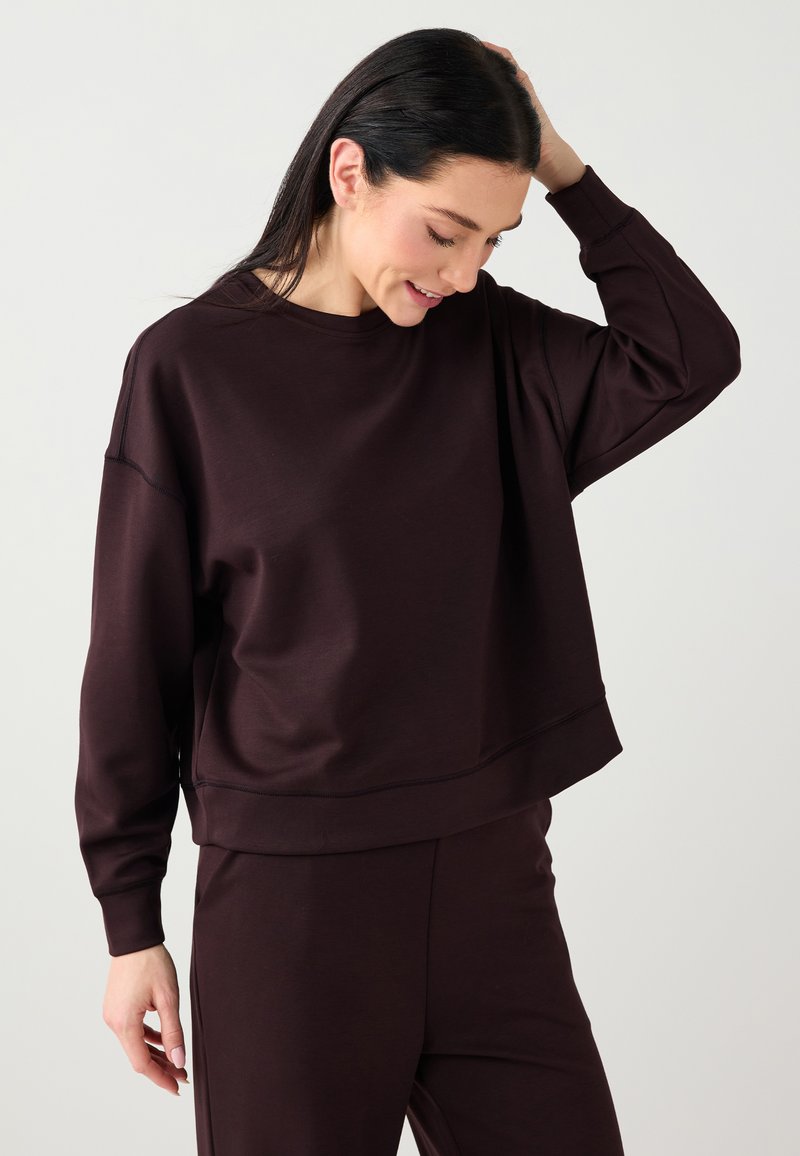 Sweat-shirt bordeaux foncé à coupe décontractée, avec poignets côtelés et encolure large. Le tissu semble lisse avec une légère brillance. Associé à un pantalon assorti.