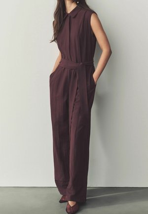 Ærmeløs mørkelilla buksedragt med krave, bundet taljebælte, vide ben og lommer, båret med matchende spidse sko.