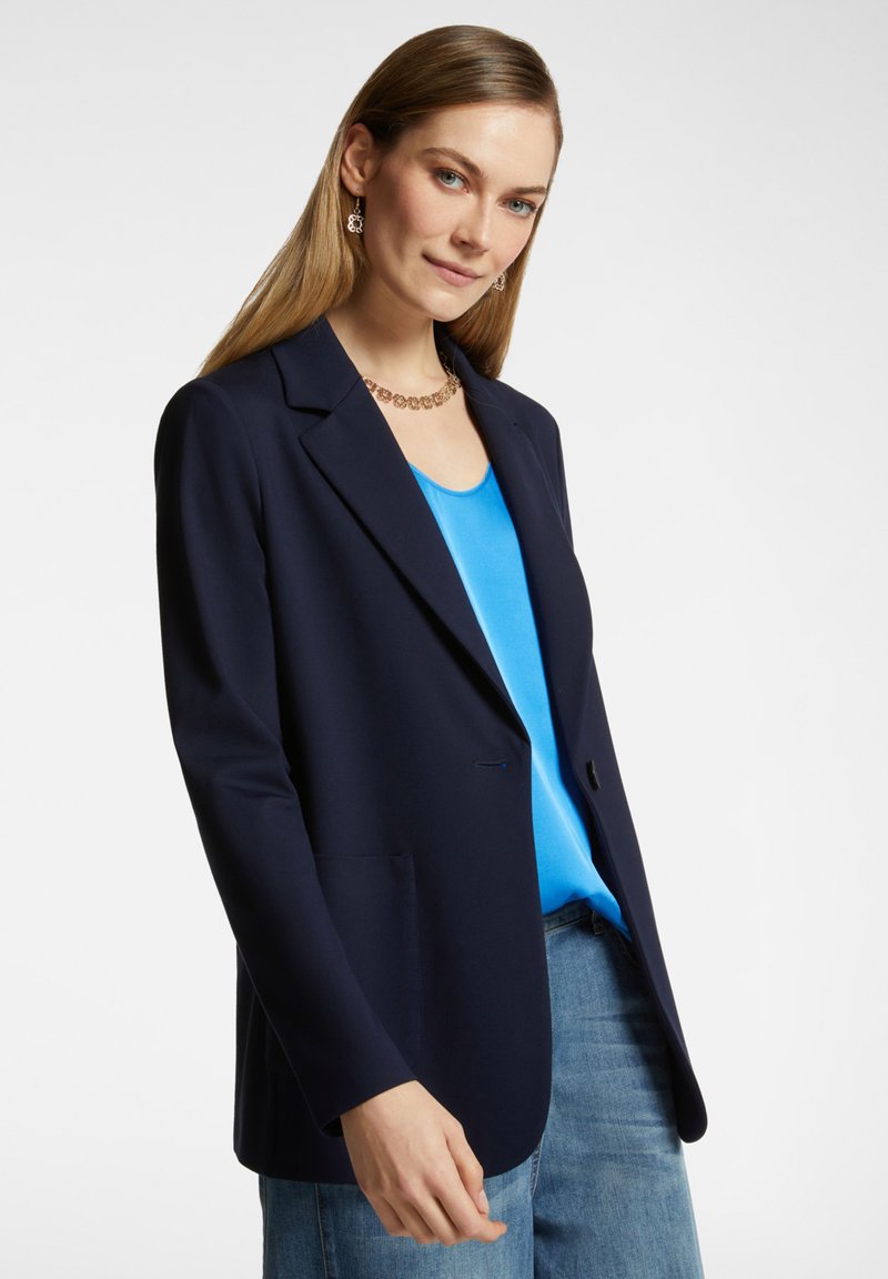 Marinblå blazer i slät tyg, med en enkel knapp, snibbkrage och två framfickor, i kombination med en ljusblå topp och jeans.