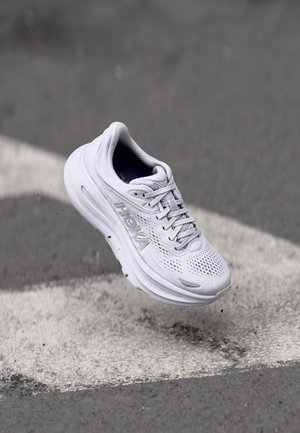 Baskets de course blanches avec une tige en mesh, une semelle épaisse et un système de laçage. Présente le logo HOKA sur le côté.