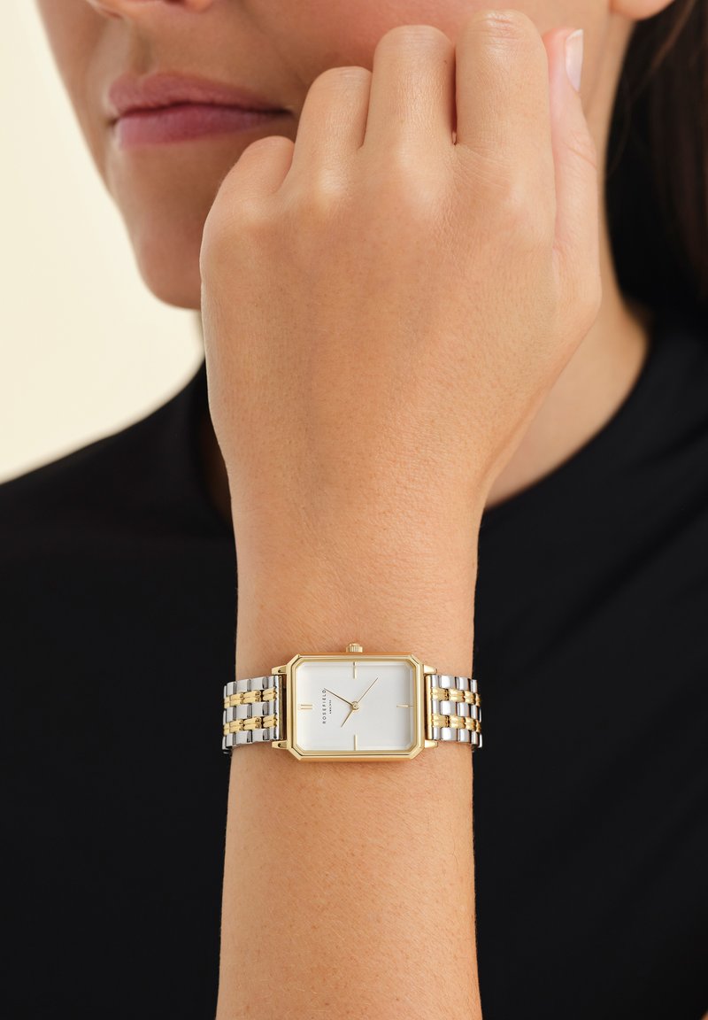 Montre rectangulaire argentée et dorée avec un cadran blanc, bracelet en métal présentant des maillons alternés, portée au poignet sur un fond noir.