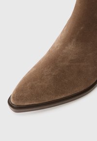 Bottine en daim brun avec un bout pointu et une texture lisse. Elle présente un design élégant et une semelle fine brune.