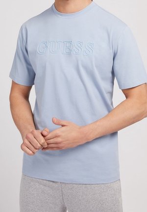 Mann, der die Hände hält, trägt ein hellblaues Kurzarm-T-Shirt mit erhabenem "GUESS"-Logo und hellgraue Jogginghose.