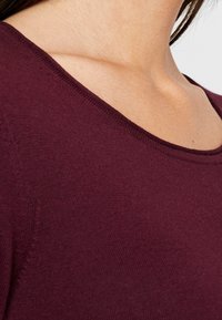 Femme portant un pull ras du cou violet foncé, laissant découvrir la clavicule et la partie supérieure de la poitrine, sur un fond neutre.