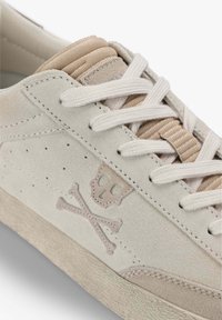 Zapatillas de ante beige con cordones blancos, que presentan un logo de calavera y tibias cruzadas. Suela texturizada y acentos lisos en la lengüeta.