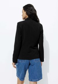Blazer noir avec une coupe structurée et des manches longues, associé à un short en denim bleu. Coutures visibles et une texture subtile sur le tissu.
