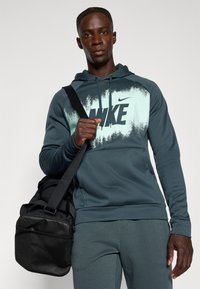 Ciemnozielona bluza Nike z logo, z efektem gradientu, połączona z dopasowanymi dresami. Czarna torba na ramię z paskiem.