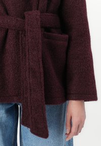 Burgundy uldblandet frakke med en tekstureret overflade, der har et bindebånd og forlommer, stylet over blå denimjeans.