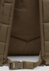 Panneau arrière de sac à dos en tissu marron avec des bretelles rembourrées et une fermeture à boucle en plastique réglable au centre en bas.