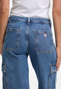 Blaue Denim-Cargohosen mit einem Lockeren Schnitt, doppelt genähten Gesäßtaschen und einem kleinen, dreieckigen Logo-Patch. Auffällige Nähte und strukturierter Stoff.