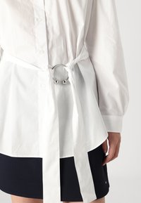 Camicia bianca in cotone con vita arricciata, dotata di accessori metallici argentati e dettaglio annodato. La camicia ha maniche lunghe e un orlo curvo.