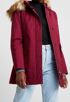 Close-up van een persoon die een bordeauxrode winterjas met bontkraag, een zwart geribd shirt en lichtblauwe jeans draagt.