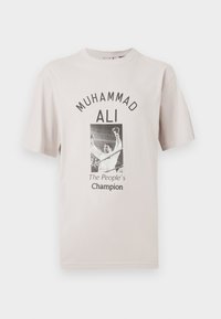 Bežs kokvilnas t-krekls ar centrālo grafiku, kurā attēlots Muhammeds Ali, teksts virs attēla lasāms "MUHAMMAD ALI," bet zem tā "Tautas čempions."