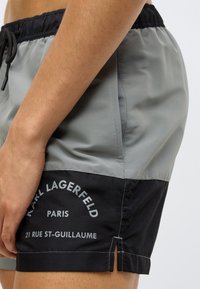 Férfi fürdőnadrág, amelynek felső fele halvány szürke, alsó fele fekete. Oldalzsebekkel, rugalmas derékrésszel és nyomtatott márkajelzéssel rendelkezik.