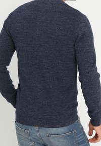 Marineblauer gestrickter Pullover mit strukturiertem Muster, gerippten Bündchen und Saum. Von hinten gezeigt, kombiniert mit hellblauen Jeans.