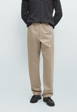 Personne portant un pantalon beige à jambes droites, des chaussures en cuir noires et une chemise noire à manches longues avec des rayures horizontales blanches.