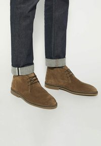 Brown suede ankle boots with a smooth texture, lace-up design, and rounded toe, paired with dark denim jeans rolled at the cuffs.
