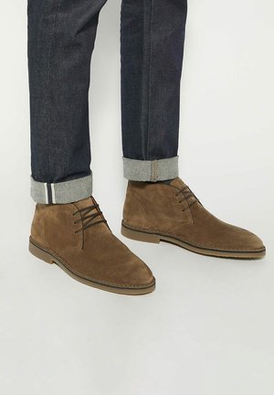 Brown suede ankle boots with a smooth texture, lace-up design, and rounded toe, paired with dark denim jeans rolled at the cuffs.