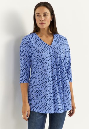 Frau trägt ein blaues V-Ausschnitt-Oberteil mit schwarzen Punkten und Dreiviertelärmeln, steht vor einem schlichten weißen Hintergrund.