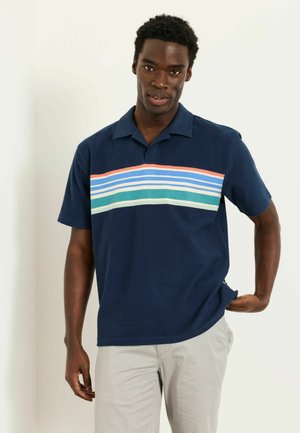 Mann trägt ein marineblaues kurzärmeliges Polohemd mit mehrfarbigen horizontalen Streifen über der Brust und hellgraue Hose.