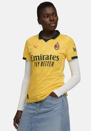 Geel voetbalshirt met een groene kraag, voorzien van het Puma-logo en "Emirates Fly Better" in het zwart. Korte mouwen, textuurontwerp.