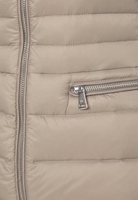 Beige vadderad jacka med quiltad design, utrustad med en metallisk dragkedjeficka och slät yta. Dragkedjan syns framtill.