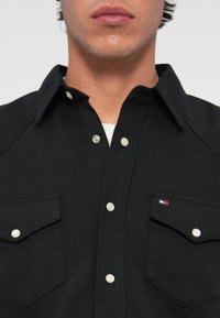 Cămașă neagră cu nasturi, realizată dintr-un material texturat, prezentând două buzunare pe piept dotate cu nasturi albi și un logo mic pe buzunarul din stânga.