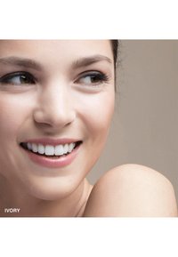 Gros plan sur un visage souriant avec une peau brun clair, des dents blanches éclatantes et un maquillage naturel. Fond flou dans des tons neutres.