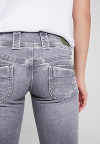 Jeans de denim gris con un acabado desgastado, que presentan dos bolsillos traseros, pasadores para cinturón y un detalle de etiqueta verde en la cintura.