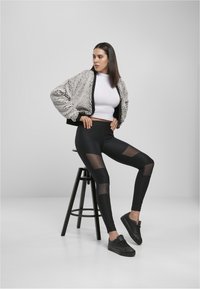 Zwarte geribbelde legging met meshpanelen, wit aansluitend topje en een grijze zachte jas, gecombineerd met zwarte sneakers, zittend op een zwarte kruk.