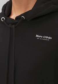 Hoodie noir en tissu doux, avec un cordon de serrage au cou et un logo blanc imprimé sur la poitrine. Design et texture simples.