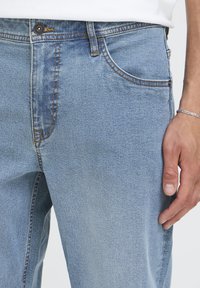 Helerohelised teksapüksid lõdva lõikega, millel on viis taskut, kontrastsed õmblused ja nööpidega esitasku, mis näitab pehmet tekstuuri.