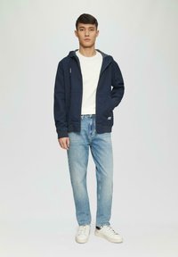 Veste à capuche légère bleu marine avec poches à l'avant, assortie à un t-shirt blanc et un jean bleu clair. Baskets blanches avec accents noirs.