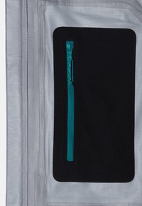 Zilveren stof met een textuurfinish, voorzien van een zwarte ritszak met een turquoise rits. Het ontwerp is stijlvol en functioneel.