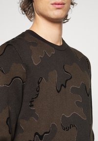 Dunkelbrauner Pullover mit Camouflage-Muster, ausgestattet mit einem Rundhalsausschnitt und einem strukturierten Strickdesign. Das "HUGO"-Logo ist dezent in das Muster integriert.