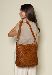 Woman with long brown hair carries large brown woven leather shoulder bag wearing beige shirt and light-striped pants.