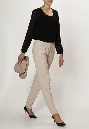 Mujer caminando con una blusa negra transparente, pantalones beige, tacones negros, sosteniendo un sombrero beige de ala ancha en su mano izquierda.