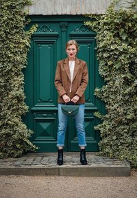 Hnědá semišová blejzr, bílá blůza s krajkovými detaily, světle modré manžestrové džíny, černé kotníkové boty, zelená kabelka, před zelenými dveřmi a pozadím s břečťanem.
