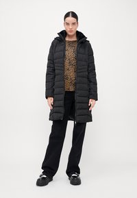 Melnā kviltotā puffer jaka ar kapuci un priekšējo rāvējslēdzēju, uzvilkta virs brūna leopardu raksta džempera. Papildināta ar melnām platajām biksēm un apaviem.