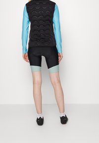 Person klädd i svarta vadderade cykelshorts och svart väst med blå långa ärmar, stående vänd mot en vit vägg och en enkel golv.