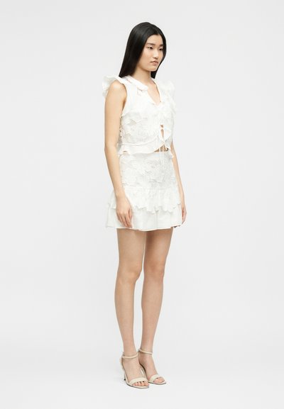 Νεαρή γυναίκα όρθια, με λευκό δαντελένιο τοπ χωρίς μανίκια και ασορτί μίνι φούστα με βολάν, φορώντας μπεζ πέδιλα με λουρί γύρω από τον αστράγαλο.