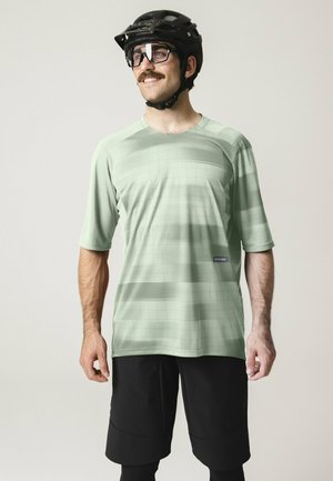 Mann trägt ein hellgrünes kurzärmeliges Radsporttrikot, schwarze Shorts, schwarzen Helm und Schutzbrille, lächelt und blickt zur Seite.