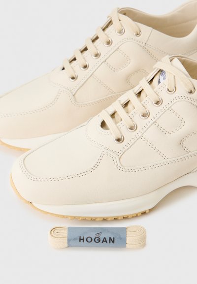 Beige ādas tenisi ar auklām un redzamām šuvēm, apvienoti ar papildu beisga auklām, uz kurām ir uzraksts "Hogan" pelēkā lentē.