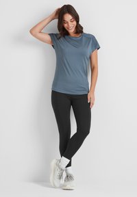 Woman standing against a white wall, wearing a blue short-sleeve shirt, black leggings, white socks, and gray running shoes, smiling and touching her hair.