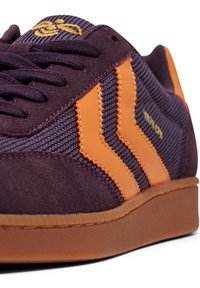 Närbild av lila sneakers med orange chevronränder, textilmaterial, mockadetaljer och brun gummisula.
